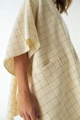 Ruana corta de lana virgen color beige con diseño a cuadros finos oscuros y bolsillos frontales aplicados.