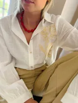 Camisa de manga larga en color blanco, confeccionada en tejido con textura. Presenta un diseño con bordado artesanal en relieve de color crema sobre uno de los hombros y parte del pecho, con cierre frontal de botones.