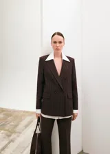 Blazer de corte sastrero en color marrón oscuro con diseño de rayas diplomáticas verticales. Presenta solapas clásicas, cierre frontal con botones y bolsillos laterales con solapa.