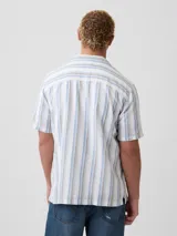 Camisa de manga corta de lino con diseño de rayas verticales en tonos blancos, azules y marrones, cuello camisero y cierre frontal de botones.