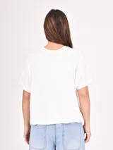 Remera de manga corta color beige claro, con cuello redondo y estampado floral en tonos dorados y plateados. Las mangas tienen un detalle de tejido acanalado.