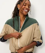 Campera rompeviento de diseño tricolor con bloques de color en verde, beige y blanco. Presenta cuello alto, cierre frontal con cremallera, mangas raglán y puños ajustados. El ruedo incluye un cordón regulable para un ajuste personalizado.
