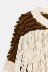 Jersey de punto con cuello redondo y manga larga, caracterizado por un diseño de parches en tonos tierra y crema. Presenta una textura variada con detalles de flecos y tejido en relieve.