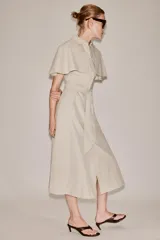 Vestido camisero midi de algodón con diseño de capa, cuello solapa, manga corta tipo capa y trabillas en los hombros. Presenta cierre frontal con botones, bolsillos delanteros y cinturón ajustable del mismo tejido con lazada. El bajo tiene un acabado en línea evasé.
