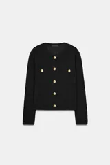 Chaqueta de punto negra, de corte cropped y manga larga, con cuello redondo. Presenta botones dorados en el frente y dos bolsillos de plastrón decorativos con botón dorado en el pecho.