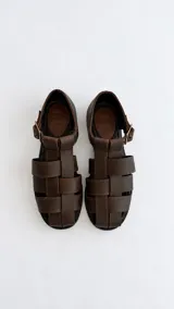 Sandalias cangrejeras de cuero marrón oscuro, con diseño de tiras entrelazadas, punta redonda, talonera cerrada y tira ajustable con hebilla metálica en el tobillo.