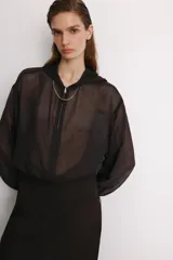 Cazadora semitransparente de color negro, con capucha, cierre frontal de cremallera metálica y mangas largas abullonadas con puños elásticos. Presenta un diseño holgado con bajo elástico.