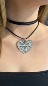 Gargantilla tipo choker de doble tira de gamuza negra con un dije metálico plateado en forma de corazón que presenta un diseño de rostro grabado.