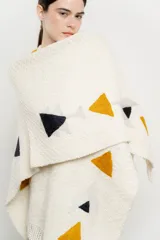 Ruana de tejido grueso color crema con flecos en el borde inferior y diseño geométrico en ocre, negro y gris.