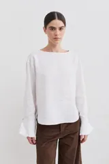 Blusa de lino con cuello bote y mangas largas terminadas en puños acampanados con volados. Presenta un cierre con botón en la espalda.