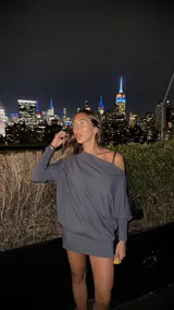 Vestido corto color gris oscuro, con escote asimétrico dejando un hombro al descubierto, mangas largas y corte holgado.