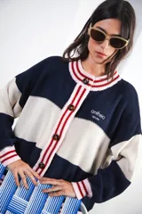 Cardigan de punto grueso con diseño a rayas horizontales en azul marino y crema. Presenta cuello redondo, cierre frontal con botones grandes y detalles de franjas en color rosa y rojo en el cuello y la botonera. Incluye el logo de la marca bordado en el pecho.