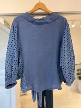 Camisa azul de manga larga con diseño abullonado, detalles de bordado estilo broderie en las mangas y laterales, cuello camisero, cierre frontal con botones y lazo para atar en el frente.