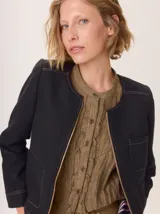 Chaqueta de paño color marrón oscuro, de corte clásico y cuello redondo, con cierre frontal dorado. Presenta bolsillos rectangulares con ribetes en color rojizo y parches de pana a rayas en los hombros.