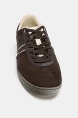 Zapatillas deportivas de gamuza en color marrón oscuro, con detalles de franjas laterales en color crema y refuerzo en el talón. Presentan cierre mediante cordones y suela de goma texturizada.