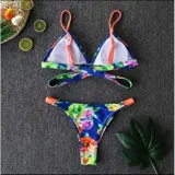 Bikini de dos piezas con estampado floral sobre fondo azul. El top es de estilo triángulo con tiras naranjas y diseño cruzado en la parte inferior del busto. La bombacha es tipo tanga con laterales regulables y detalles en color naranja.