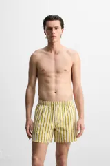 Short de baño confeccionado en tejido técnico, con diseño de rayas verticales amarillas y verdes sobre fondo blanco. Cuenta con cintura elástica ajustable con cordón, bolsillos laterales y bolsillo trasero con vivo.