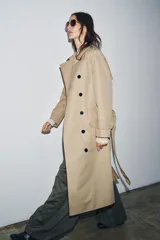 Trench color beige con cuello solapa y manga larga ajustable con trabillas. Bolsillos en delantero. Cinturón en mismo tejido. Detalle de pespuntes marcados a tono. Cierre frontal cruzado con botones. Tejido repelente al agua.