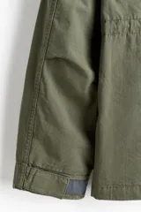Campera estilo cargo de tejido ripstop, color verde militar, con cuello elevado que incluye capucha oculta. Presenta cierre frontal con solapa rompevientos, cuatro bolsillos delanteros con solapa y botón a presión, puños ajustables y cordón interno en la cintura.