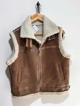 Chaleco estilo gamulán con interior y bordes de corderito sintético. Presenta cierre frontal, cuello solapa con hebillas decorativas y bolsillos laterales.