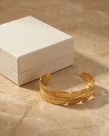 Pulsera tipo esclava rígida de acero con baño de oro, diseñada con una textura detallada que simula una pluma envolvente.