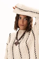 Buzo de tejido de punto color crudo con diseño de trenzas y costuras decorativas en color negro que recorren la prenda en forma de espiral. Presenta cuello redondo y mangas largas con terminación acampanada.