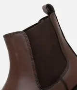 Bota tipo Chelsea de caña media, confeccionada en cuero sintético color marrón oscuro. Presenta punta redonda, paneles laterales elásticos para facilitar el calce y suela de goma robusta.