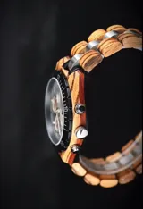 Reloj cronógrafo con caja de madera de zebrano y bisel técnico de acero negro. La esfera es plateada con subdiales de madera, indicador de fase lunar y fechador. La malla combina eslabones de acero inoxidable con madera de zebrano.