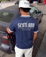 Camiseta de fútbol de Escocia de manga corta, color azul marino con diseño a cuadros sutiles, cuello en V y cuello camisero blanco con líneas azules. Presenta detalles estampados en el frente, incluyendo el escudo de la Asociación de Fútbol de Escocia, un león rampante y el texto 'Scottish Football Association'.
