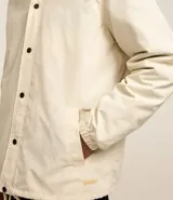 Campera color crema de poliéster con cuello inglés, abotonadura delantera con botón a presión, mangas largas con puño elástico, bajo elástico y cordón para ajustar.