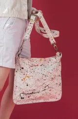 Cartera bandolera de cuero color blanco con diseño artístico de salpicaduras de pintura multicolor, correa ajustable y herrajes metálicos.