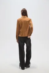 Blusa de gamuza sintética color camel, con cuello camisero y cierre frontal mediante cordones entrelazados. Presenta mangas largas y un corte holgado.