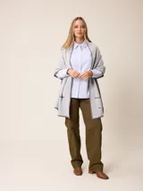 Poncho abierto de tejido de punto en color gris claro con bordes y detalles de bolsillos en gris oscuro. Presenta un diseño holgado y versátil con placa metálica decorativa en uno de los bolsillos frontales.