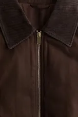 Chaleco de lona de algodón con terminación encerada en color marrón, levemente acolchado. Presenta un cuello de pana a tono, cierre frontal de cremallera, bolsillos laterales insertados y cintura elástica con frunces.