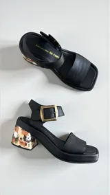 Sandalias de cuero negro Carolina De Cunto, con taco bloque medio intervenido con diseño artístico y hebilla metálica grande en tono oro viejo con logo grabado.