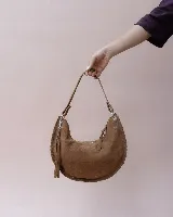 Bolso tipo hobo de gamuza color marrón, con diseño de media luna y detalles de costuras trenzadas en los bordes. Incluye una correa ajustable, cierre superior con tiradores de cuero y un detalle de flecos laterales.