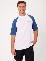 Remera holgada Rusty modelo Isel, de color blanco con mangas raglán en color azul, cuello redondo ancho y pequeño logo estampado en el pecho.