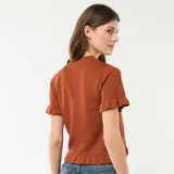Camiseta de punto color marrón, de manga corta con terminación en volantes, cuello redondo y cierre frontal con botones, decorada con una hilera de volantes a lo largo de la botonadura.
