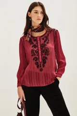 Camisa de manga larga en color negro con tejido semitransparente y detalles de hilos metalizados. Presenta un diseño con bordados florales en tonos tierra, cierre frontal con botones y un lazo decorativo en el cuello.