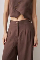 Pantalón de tiro alto color chocolate, con pinzas frontales que aportan caída suave y silueta amplia de pierna recta. Cierra con botón y bragueta, y suma un detalle de botón funcional en la cintura que le da un aire sastrero.