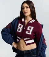 Cardigan de punto estilo college color bordó, con cierre frontal de doble cremallera, mangas largas y detalles de rayas blancas en cuello, puños y ruedo. Presenta una estampa numérica blanca en el frente.