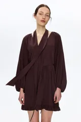 Vestido oversized corto de color marrón oscuro, con escote en V pronunciado y pañuelo haciendo juego que puede abrocharse en la nuca. Cuenta con botones forrados adelante, cinturón fino para atar, mangas globo largas de corte raglán con elástico en los puños y ribete de volados en el bajo.