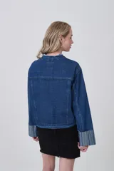 Campera corta de denim azul oscuro, con cuello mao deshilachado y botones plateados frontales. Las mangas largas tienen puños anchos con estampado de rayas verticales en tonos celestes y blancos.