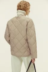 Campera acolchada de color beige con diseño guateado en rombos, hombros caídos, cuello redondo, cierre frontal de botones y bolsillos parche delanteros. Incluye una bufanda a juego que se abrocha en la nuca y cuenta con aislamiento térmico THERMOLITE.