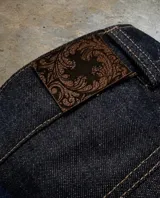 Bermuda de jean estilo raw denim, confeccionada en algodón de 12 onzas. Presenta un parche de cuero grabado con diseño barroco en la parte posterior y costuras reforzadas en contraste.