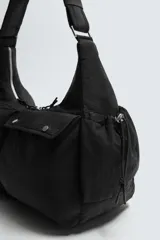 Bolso de hombro negro, fabricado en tejido técnico, con múltiples bolsillos exteriores, incluyendo uno con cierre de cremallera y otro lateral. El compartimento principal y un bolsillo interior también tienen cierre de cremallera. Cuenta con un asa de hombro ajustable.