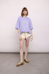 Remera lila de corte holgado con mangas cortas y estampado circular con figuras de nadadoras y texto "The Ped Swim Club".