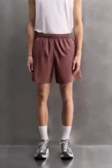 Pantalones cortos deportivos color marrón rojizo, confeccionados en tejido técnico ligero y elástico. Presentan cintura elástica ajustable con cordón interior, bolsillos frontales y un bolsillo trasero con cierre de cremallera. Los laterales tienen perforaciones para transpirabilidad y aberturas laterales en el bajo.