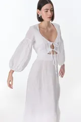 Vestido largo blanco de lino, con escote en pico, mangas largas abullonadas y lazos frontales. Presenta tajos en la falda.