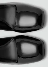 Zapato de tacón alto y ancho, color negro con acabado charol, punta cuadrada y tira ajustable con hebilla al tobillo.